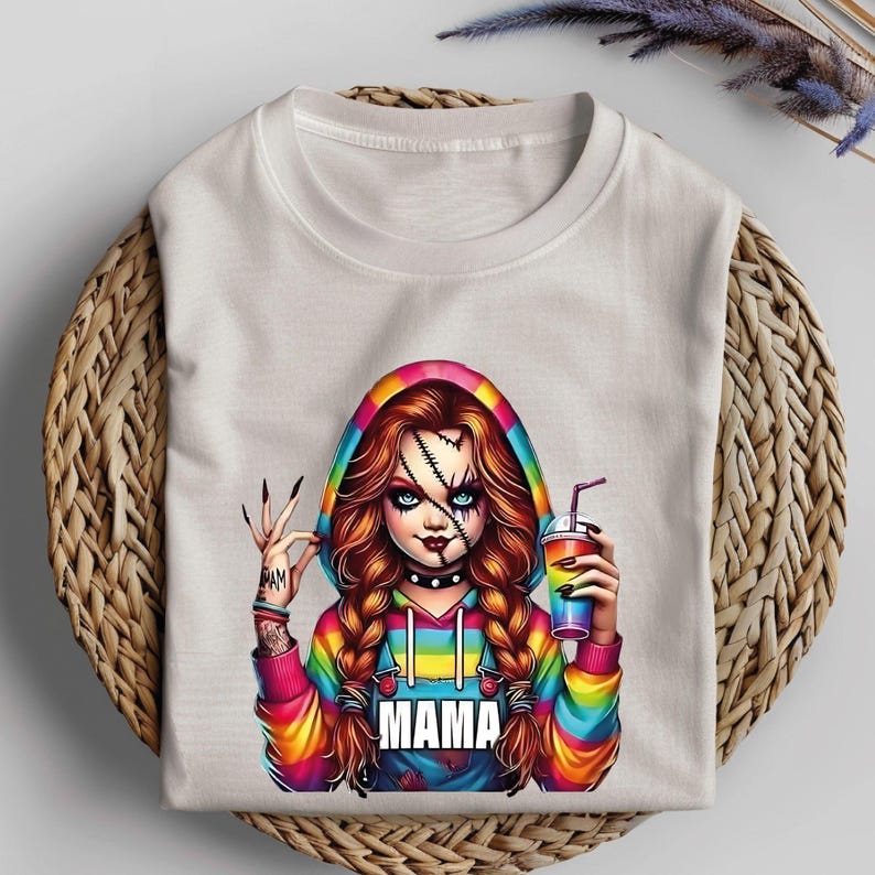 May include: White t-shirt featuring a colorful graphic of a doll-like character with stitched facial details, holding a drink. The character wears a rainbow hoodie with the word "MAMA" on the front. The character has long, braided hair and a tattoo on the arm.