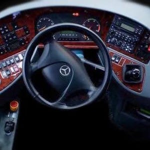 Op de afbeelding: De cockpit van een bus met een zwart stuurwiel voorzien van een Mercedes-Benz logo, een dashboard met houtnerfpatroon, diverse bedieningsknoppen, een digitaal instrumentenpaneel en een versnellingspook aan de rechterkant.