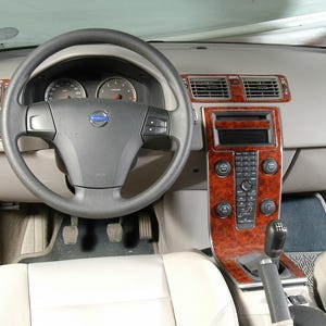 Könnte beinhalten: Innenansicht eines Volvo-Autodashboards. Das Armaturenbrett ist hellgrau mit Holzmaserungsakzenten. Das Lenkrad ist schwarz mit dem Volvo-Logo in der Mitte. Die Mittelkonsole verfügt über ein Radio und Klimaanlagen.