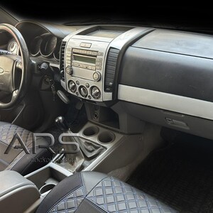 Peut inclure: L'intérieur d'un véhicule avec un volant noir, une console centrale argentée dotée de commandes audio et des sièges sombres avec un motif en losanges bleus. Le tableau de bord comprend des bouches d'aération circulaires et un levier de vitesse.