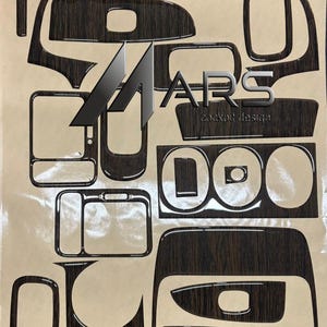 Può includere: Un set di adesivi per interni con motivo venatura del legno marrone scuro per l'abitacolo di un veicolo. I pezzi adesivi presentano varie forme geometriche per il cruscotto, con il testo MARS cockpit design stampato al centro.