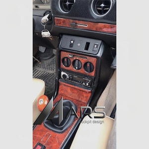 Puede incluir: Una consola central de vehículo con acabado de madera, rejillas de ventilación negras, radio digital, tres perillas circulares de control de clima y una funda de palanca de cambios negra. Un llavero blanco con forma de coche cuelga del encendido.