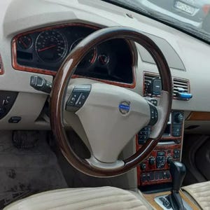 Op de afbeelding: Interieur van een Volvo-auto. Het dashboard is beige met houten accenten. Het stuurwiel is bruin met beige bedieningselementen en een Volvo-embleem. Het interieur van de auto heeft een snelheidsmeter, ventilatieopeningen en een versnellingspook.
