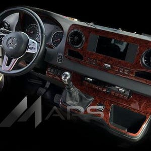 May include: A detailed shot of a Mercedes-Benz vehicle's dashboard. The interior features a steering wheel, instrument panel, and a central console with a screen. The trim has a wood grain effect, and the color palette includes brown, black, and grey.