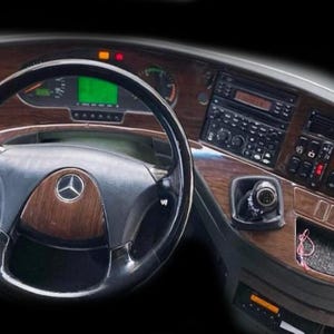 Op de afbeelding: Een voertuigdashboard met een houtnerfafwerking, een zwart stuurwiel met een centraal Mercedes-Benz-logo, een digitaal instrumentenpaneel, een radio-eenheid en diverse bedieningsknoppen en schakelaars op de console.
