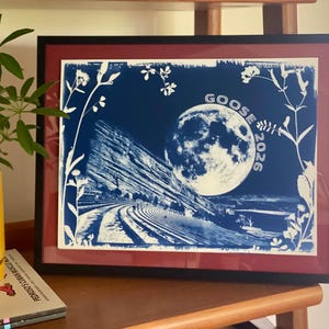 May include: A framed cyanotype print featuring a blue and white landscape of a large rock formation and a full moon, with the text GOOSE 2026 printed on the right side. The image is bordered by white silhouettes of various wildflowers and plants.