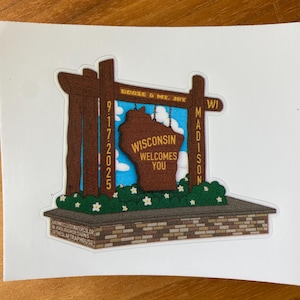 May include: A sticker featuring a brown sign that reads "Wisconsin Welcomes You" in front of a blue sky background. The sign is framed by brown posts with the text "Goose & Mt. Joy" and "Madison WI". The sticker also includes the date 9/17/2025.