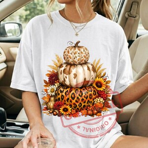 Könnte beinhalten: Ein weißes Kurzarm-T-Shirt mit einer Grafik aus drei gestapelten Kürbissen. Der oberste Kürbis hat ein Eichenblattmuster, der mittlere ein Kuhfellmuster und der untere ein Leopardenmuster, umgeben von Sonnenblumen und herbstlichen Blättern.