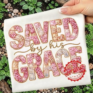 Op de afbeelding: Wit t-shirt met de tekst "SAVED by his GRACE" in glitterende roze, gouden en zilveren letters. De woorden zijn goud omrand. Een rode "BEST SELLER" stempel staat rechtsonder.