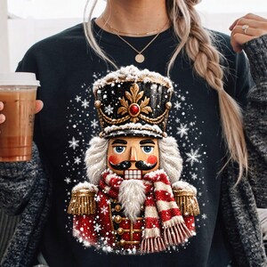 Puede incluir: Una sudadera negra de manga larga con un estampado gráfico de un soldado cascanueces que lleva un sombrero negro y dorado, una chaqueta roja con adornos dorados y una bufanda a rayas rojas y blancas, rodeado de copos de nieve blancos.