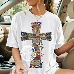 May include: White t-shirt featuring a large cross design. The cross is composed of camouflage patterns and American flag elements. The words "God Bless America" are printed vertically on the right side of the cross. A "Best Seller" sticker is at the bottom.