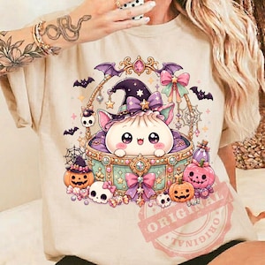 May include: A beige t-shirt featuring a graphic of a white dumpling character wearing a purple witch hat inside an ornate, decorated basket. The design includes pink pumpkins, small skulls, spider webs, and bows in shades of purple and teal.