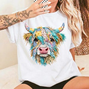 Könnte beinhalten: Weißes T-Shirt mit einer farbenfrohen Patchwork-Illustration eines Highland-Kuhgesichts. Das Fell der Kuh ist eine Mischung aus Blau, Gelb und Grün, mit gemusterten Stoffakzenten auf den Hörnern und im Gesicht. Die Kuh hat eine rosa Nase und dunkle Augen.