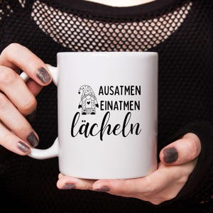 Może przedstawiać: Biały ceramiczny kubek z czarno-białą ilustracją gnoma i napisami "AUSATMEN, EINATMEN, l&auml;cheln". Kubek ma biały uchwyt i jest trzymany przez osobę z ciemnym lakierem do paznokci.
