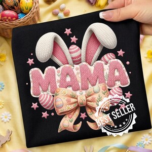 Puede incluir: Camiseta negra con un diseño "MAMA" rosa tejido, orejas de conejo, huevos de Pascua y un lazo. El diseño incluye estrellas rosas y un sello de "Mejor vendedor". Ideal para Pascua o primavera.
