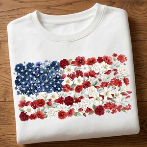 Könnte beinhalten: Weißes Sweatshirt mit einem floralen amerikanischen Flaggen-Design. Die Sterne der Flagge sind weiß, und die Streifen bestehen aus roten, weißen und blauen Blumen, darunter Rosen, Mohnblumen und Gänseblümchen. Ein patriotisches Thema.