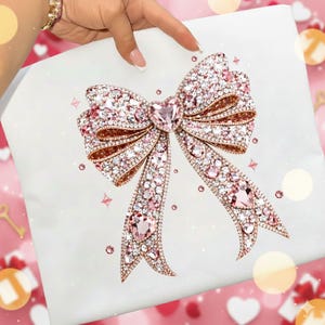 Puede incluir: Un lazo decorativo con un acabado metálico oro rosa e incrustado con cristales rosas y transparentes. El lazo está centrado en una superficie blanca, con un cristal en forma de corazón en el centro. El diseño del lazo incluye múltiples bucles y colas fluidas.