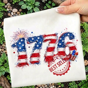 Op de afbeelding: Wit t-shirt met het nummer "1776" in een patriottisch ontwerp met de kleuren van de Amerikaanse vlag. Het ontwerp bevat vuurwerk, sterren en een "Best Seller" zegel, perfect voor Independence Day vieringen.