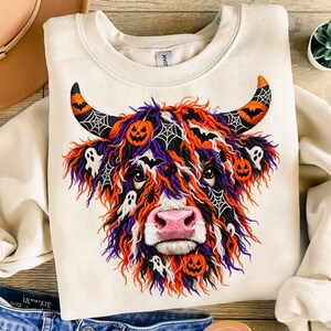 Könnte beinhalten: Ein cremefarbenes Sweatshirt mit Rundhalsausschnitt und einem Grafikdruck eines Hochlandrindes mit langem, zotteligem Fell in Orange, Violett und Weiß. Das Rind ist mit Halloween-Motiven wie Kürbissen, Geistern, Fledermäusen und Spinnennetzen verziert.