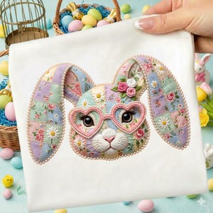 Könnte beinhalten: Ein weißes quadratisches Stoffstück mit einem gestickten Hasengesicht. Der Hase hat Flickenohren, herzförmige Brille und ist mit Blumen und Perlen verziert. Das Design ist in Pastellfarben gehalten, perfekt für Ostern.