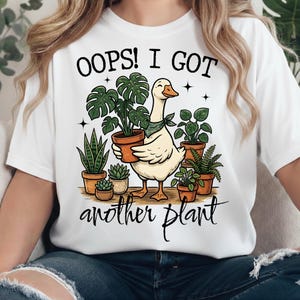 Puede incluir: Camiseta blanca con un dibujo de un ganso de dibujos animados sosteniendo una planta en maceta, rodeado de otras plantas en maceta. El texto de la camiseta dice "OOPS! I GOT another plant."