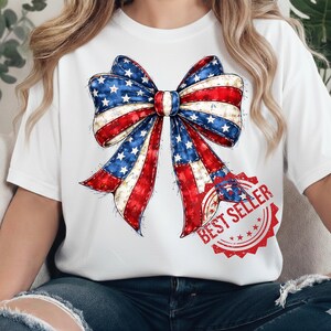 Op de afbeelding: Wit T-shirt met een groot strikdesign in rood, wit en blauw, dat doet denken aan de Amerikaanse vlag. De strik heeft een distressed, vintage uitstraling. Een rode "BEST SELLER" stempel staat rechtsonder.