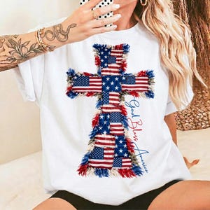 Puede incluir: Camiseta blanca con un diseño de cruz hecho con la bandera estadounidense. La cruz está delineada con flecos rojos, blancos y azules. Las palabras "God Bless America" están escritas en azul en el lado derecho de la cruz.