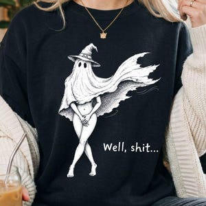 May include: A black crewneck sweatshirt featuring a white line art illustration of a figure draped in a sheet wearing a witch hat, with the text Well, shit... printed in white. The figure has legs exposed and is standing in a crossed-leg pose.