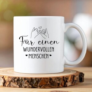 Może przedstawiać: Biały ceramiczny kubek z czarną grafiką przedstawiającą splecione dłonie z sercem nad nimi. Kubek ma niemiecki napis "F&uuml;r einen wundervollen Menschen" i dwa symbole serca. Kubek stoi na drewnianej podstawce.