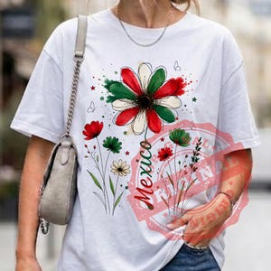 May include: A white short-sleeve t-shirt featuring a graphic of a large daisy with red, white, and green petals. The design includes smaller wildflowers, butterflies, and the word Mexico written in a cursive script font in red and green ink.