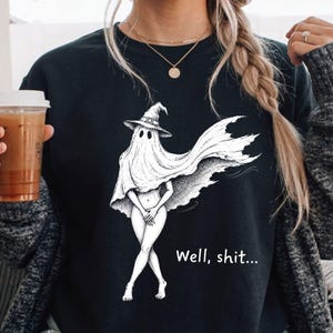 May include: A black crewneck sweatshirt featuring a white graphic illustration of a ghost wearing a witch hat and a sheet, with the text "Well, shit..." printed in white lettering below the figure. The design is centered on the front of the garment.