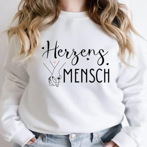 Może przedstawiać: Biała bluza z niemieckim napisem "Herzensmensch" w czarnym skrypcie, co oznacza "bratnia dusza". Projekt zawiera rysunek dw&oacute;ch trzymających się rąk z małym r&oacute;żowym sercem między nimi.