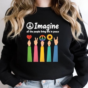 Puede incluir: Sudadera negra con la palabra "Imagine" y el símbolo de la paz. Debajo del texto hay manos coloridas que sostienen un corazón rojo, un signo de la paz, un símbolo de la paz arcoíris, un girasol y un gesto de la mano de rock. El texto dice "all the people living life in peace."