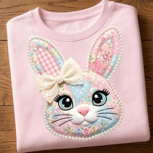May include: A pale pink sweatshirt with a decorative bunny face. The bunny has a patchwork design with floral and gingham patterns, a cream lace bow, and is outlined with faux pearls. The bunny has large eyes and a pink nose.