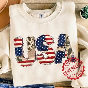 Può includere: Felpa color crema con la scritta "USA" applicata sul davanti. Le lettere sono realizzate in tessuto con motivi a bandiera americana e mucca. Un adesivo rosso "Best Seller" si trova nell'angolo in basso a destra.