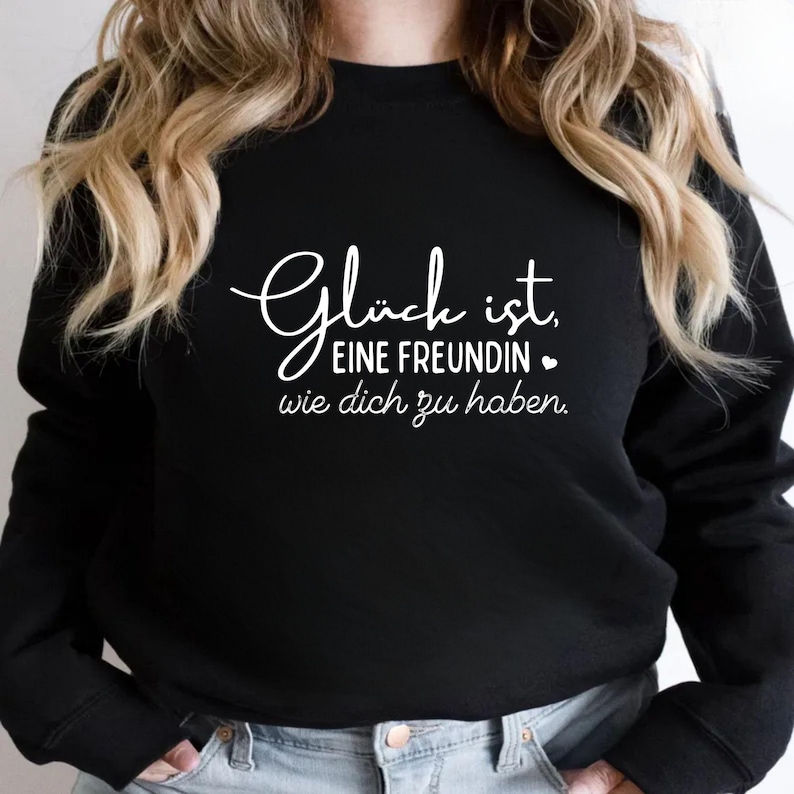 Może przedstawiać: Czarna bluza z białym napisem: "Gl&uuml;ck ist, EINE FREUNDIN wie dich zu haben." Tekst zawiera mały symbol serca. Bluza ma swobodny styl.