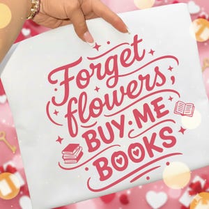 May include: White t-shirt with the text "Forget flowers; BUY ME BOOKS" in a cursive, red font. The design includes illustrations of books and hearts. The shirt is held by a hand with a gold bracelet.