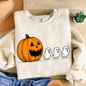 Pode incluir: Camisola creme com uma abóbora laranja esculpida e três fantasmas brancos. O design é temático de Halloween, perfeito para a época de outono e celebrações assustadoras.