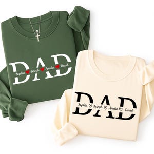 Puede incluir: Dos sudaderas de manga larga, una verde y otra crema, cada una con la palabra "DAD" en letras blancas grandes. Debajo, los nombres Sophia, Joseph, Amelia y David. Un collar de cruz plateada en la sudadera verde.