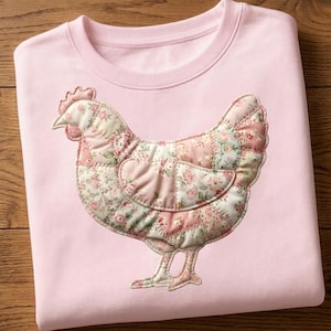 May include: A light pink sweatshirt featuring a patchwork chicken design. The chicken is made of floral patterned fabric in shades of pink, white, and green. The sweatshirt has a crew neck and long sleeves.