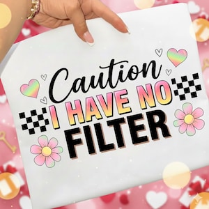 May include: White t-shirt with the text "Caution I Have No Filter" in black, yellow, and pink. The design includes hearts, flowers, and a checkered pattern. The shirt is held by a hand.
