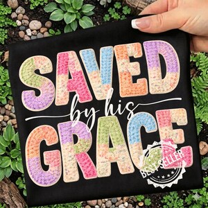 May include: Black t-shirt with the words "SAVED by his GRACE" in colorful, patchwork-style letters. The design includes a "Best Seller" stamp. The shirt is held by a hand.