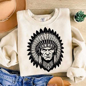 Puede incluir: Sudadera color crema con un gráfico negro de un jefe nativo americano con un tocado de plumas. El diseño está centrado en la parte delantera de la parte superior de cuello redondo y manga larga. La sudadera es de material suave.