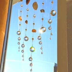 Könnte beinhalten: Dekorative Fensterdekoration mit klaren Kristallkugeln und bernsteinfarbenen Akzenten. Die Kugeln hängen an filigranen Ketten und reflektieren das Sonnenlicht vor einem blauen Himmel. Enthält natürliche, muschelartige Elemente.