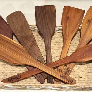 May include: A collection of wooden spatulas in various shades of brown, arranged in a woven basket. Each spatula has a flat, rectangular head and a long handle. Some handles have the word "Afterlife" engraved on them. The spatulas are made of different types of wood.
