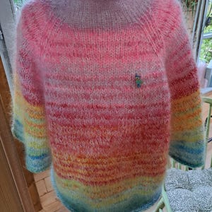 Op de afbeelding: Een handgebreide trui in een levendige kleurovergang van roze naar oranje, geel en blauw. De trui heeft een hoge, geribbelde kraag en lange mouwen. Een kleine decoratieve speld is aan de voorkant bevestigd. De trui wordt op een paspop gepresenteerd.