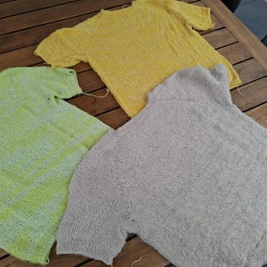 Op de afbeelding: Drie gebreide shirts met korte mouwen in geel, groen en grijs liggen op een houten tafel. De handgemaakte shirts hebben zichtbare steekpatronen en losse draden. De kleuren zijn zacht en gedempt, wat wijst op een natuurlijke vezel.