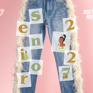 May include: Light blue denim jeans featuring white feather trim along the outer seams. The jeans are decorated with glittery green and gold letters spelling SENIOR and the numbers 2027, interspersed with illustrations of a princess character.