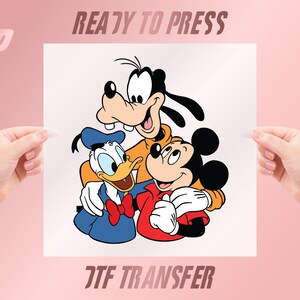 May include: A white ready-to-press DTF transfer featuring Mickey Mouse, Donald Duck, and Goofy embracing. Mickey wears a red shirt, Donald a blue sailor suit, and Goofy an orange sweater. The text "READY TO PRESS" and "DTF TRANSFER" are visible.