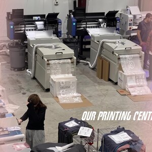 May include: A printing center featuring two large industrial printers with white output sheets, a person standing near the machines, and another person working at a station with stacks of folded garments and the text OUR PRINTING CENTER.